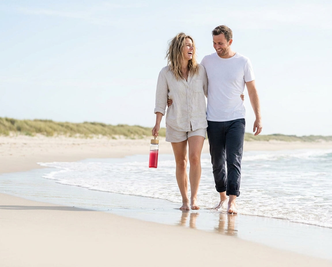 A couple walking barefoot along the shoreline, laughing, with a glass bottle of pink-red Molo in her hand